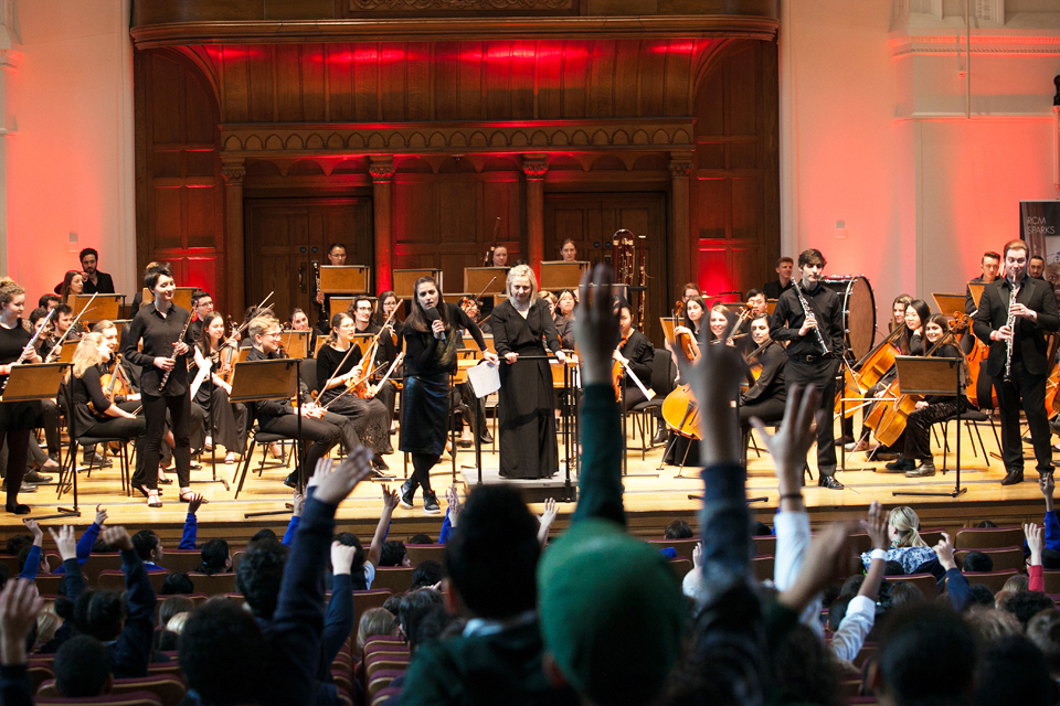 An orchestra sitting on stage with their instruments in the background, with a female presenter and   a female conductor in a conductors podium, with a group of young students putting their hands up.